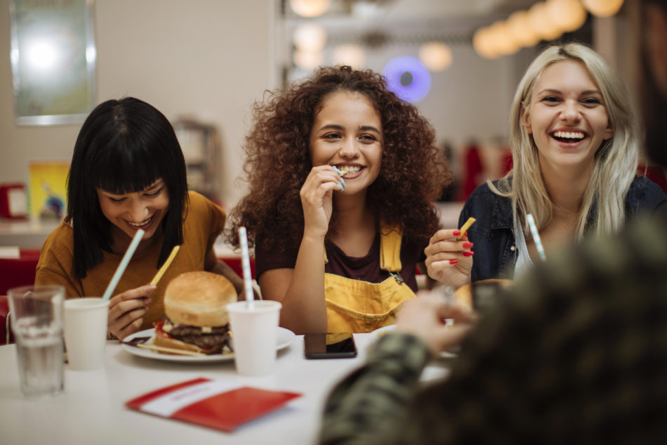female friends laughing while eating burgers and fries in a restaurant