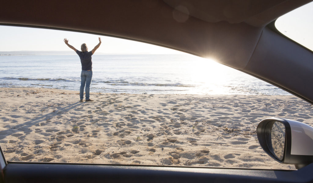 Man stretches outside car on beach, by sea shore
